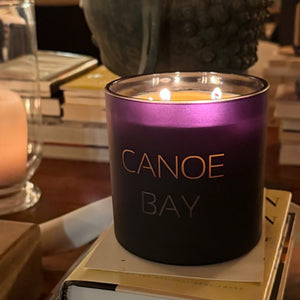 A lit candle in a BLACK glass jar with the words 'CANOE BAY' printed on the front, resting on a stack of books in a cozy indoor setting.