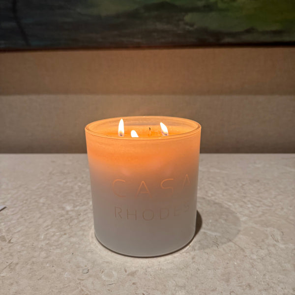 A lit Casa Rhode candle in a frosted glass jar, casting a warm orange glow on a light stone surface with a blurred background.