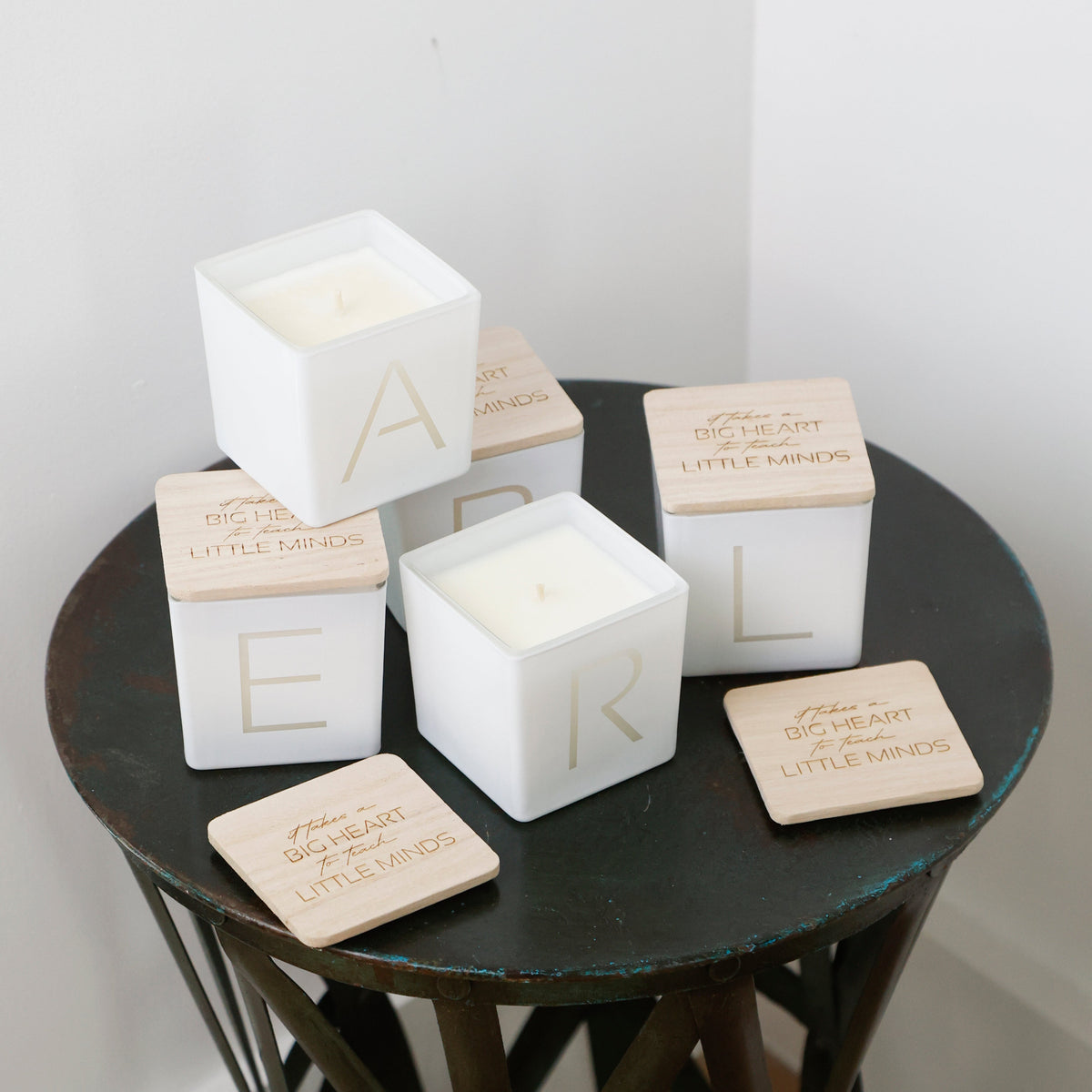 Three white square candle jars with light wood lids stacked on a dark round table, alongside matching wooden coasters. The lids are engraved with the name 'EARL' and a quote from 'The Little Things'.