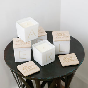 Three white square candle jars with light wood lids stacked on a dark round table, alongside matching wooden coasters. The lids are engraved with the name 'EARL' and a quote from 'The Little Things'.
