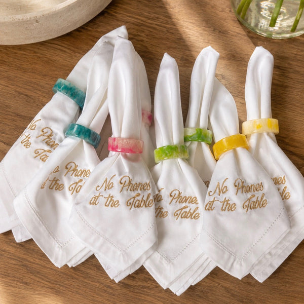 Four white napkins with gold embroidered table rules, each tied with a different colored ring and displayed on a wooden table.