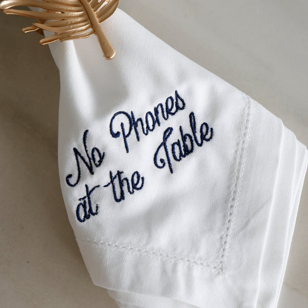 A folded white cloth napkin with navy blue embroidered text reading 'No Phones at the Table,' hanging from a gold wire ring on a light neutral surface.