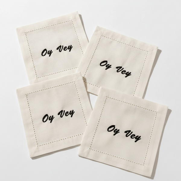 Four square white linen napkins with black embroidered script reading 'Oz Vey' inside a dashed border, arranged on a marble table with warm sunlight and shadows. Cocktail glasses and olives are partially visible around the edges.