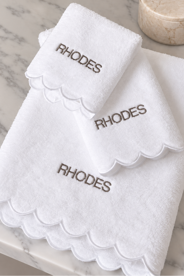 Three white towels with scalloped edges and embroidered 'RHODES' text, arranged on a light marble surface with a round stone object in the background.