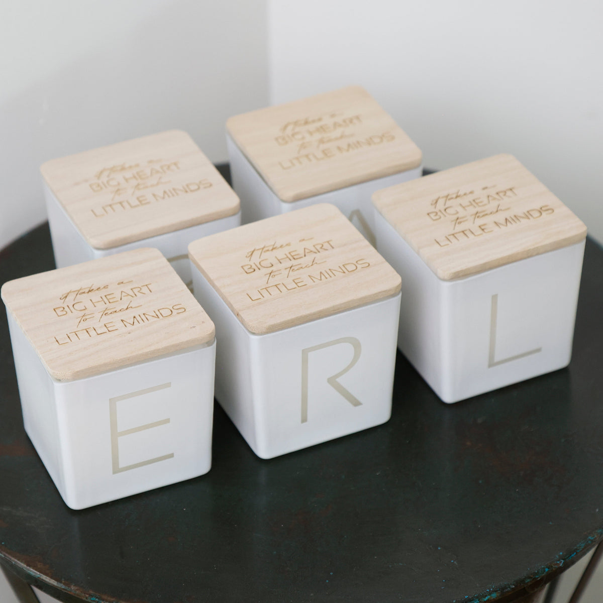 Five white square decorative boxes with light wood lids arranged on a dark round table. Each lid is engraved with the phrase 'BIG HEART LITTLE MINDS' and one box shows a large gold letter, with the others showing partial letters.
