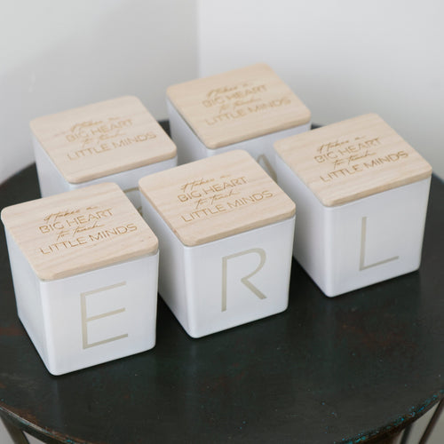 Five white square decorative boxes with light wood lids arranged on a dark round table. Each lid is engraved with the phrase 'BIG HEART LITTLE MINDS' and one box shows a large gold letter, with the others showing partial letters.