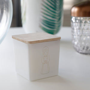 A small square white glass container with a light natural wood lid, featuring a simple line drawing of a robed figure on the front. The container sits on a white tabletop with softly blurred decorative objects in the background.