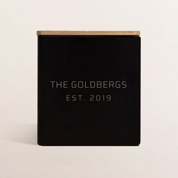 Black candle engraved The Goldbergs, Est. 2019, with a wooden lid, on a bone backdrop