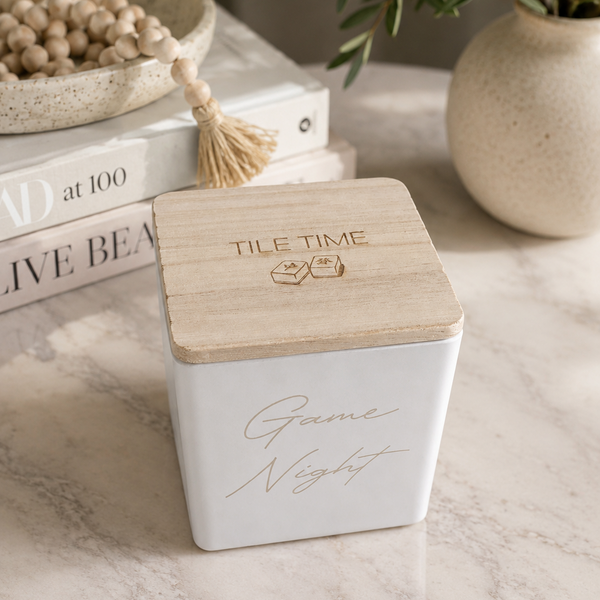 A square white glass jar engraved with "game night" with a light natural wood lid, featuring the words 'TILE TIME'. The candle is styled on a marble tabletop with books, a bowl of beads, and a potted plant in the background.