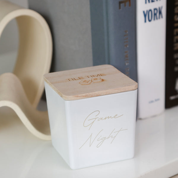 A white square candle tin with a light wood lid engraved with 'FILE FIRE' and gold script reading 'Game Night' on the front, styled on a white shelf next to books and a beige sculptural decor piece.