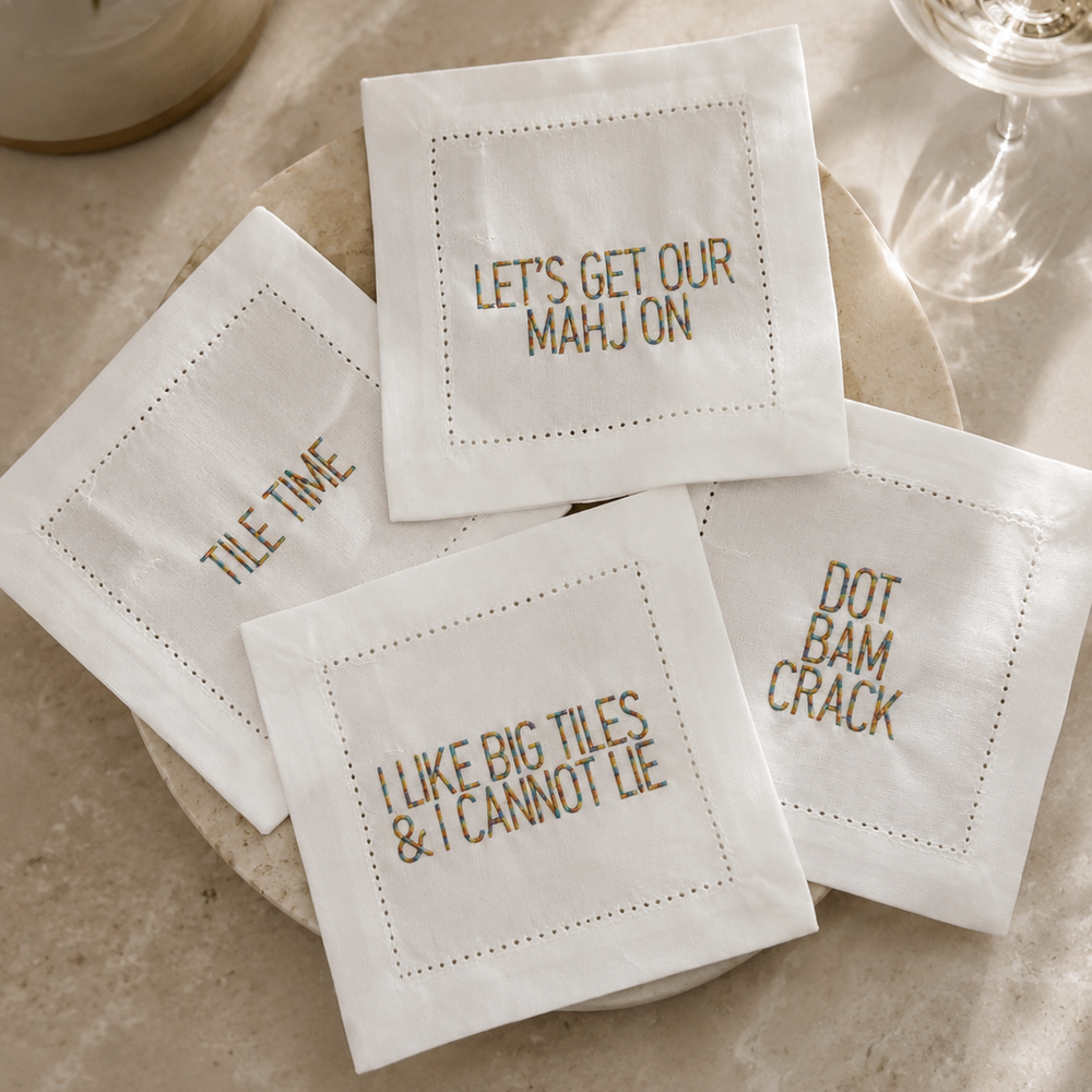 Four white square napkins with embroidered dotted borders and humorous text, arranged on a beige surface with a glass and ceramic decor in the background.