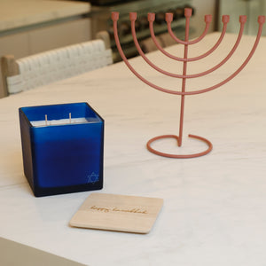 A blue glass candle holder with white wax and a Star of David, placed on a white marble kitchen island next to a pink metal menorah and a wooden 'Happy Hanukkah' coaster.