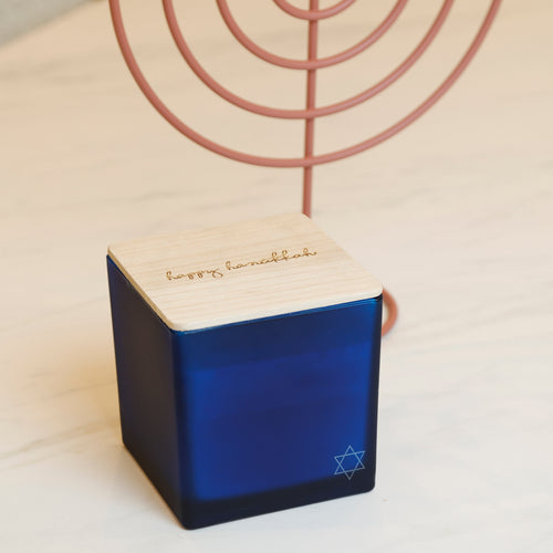 A blue and light wood Hanukkah candle holder with a rose gold metal menorah behind it, set on a light-colored wooden table.