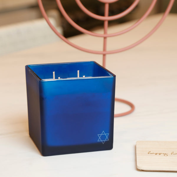 A square cobalt blue glass candle with a white Star of David logo on the bottom right, set on a light wooden table with a pink metal menorah in the background.