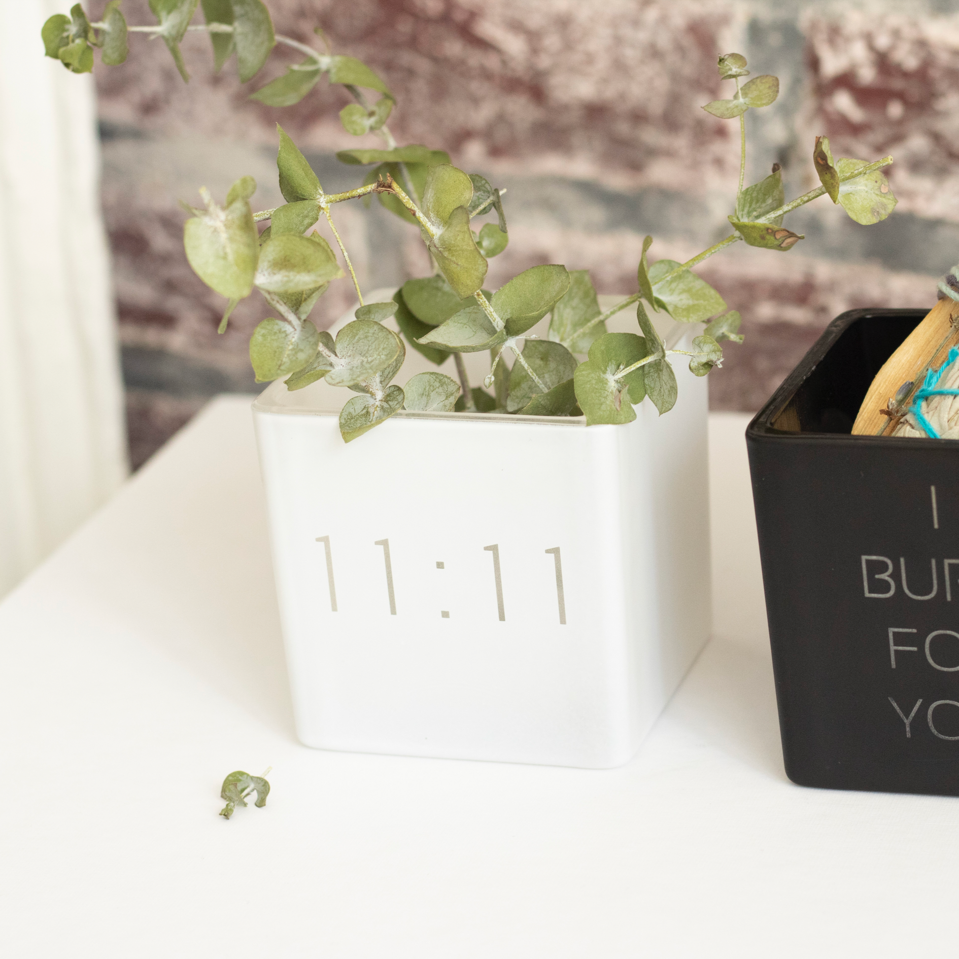 white vase with 11:11 engraved on the front, filled with eucalyptus plants. next to it black smudge kit i burn for you