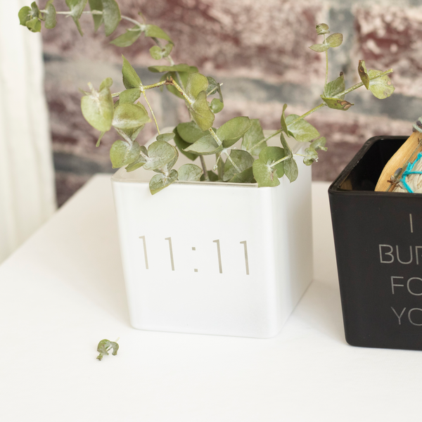 white vase with 11:11 engraved on the front, filled with eucalyptus plants. next to it black smudge kit i burn for you