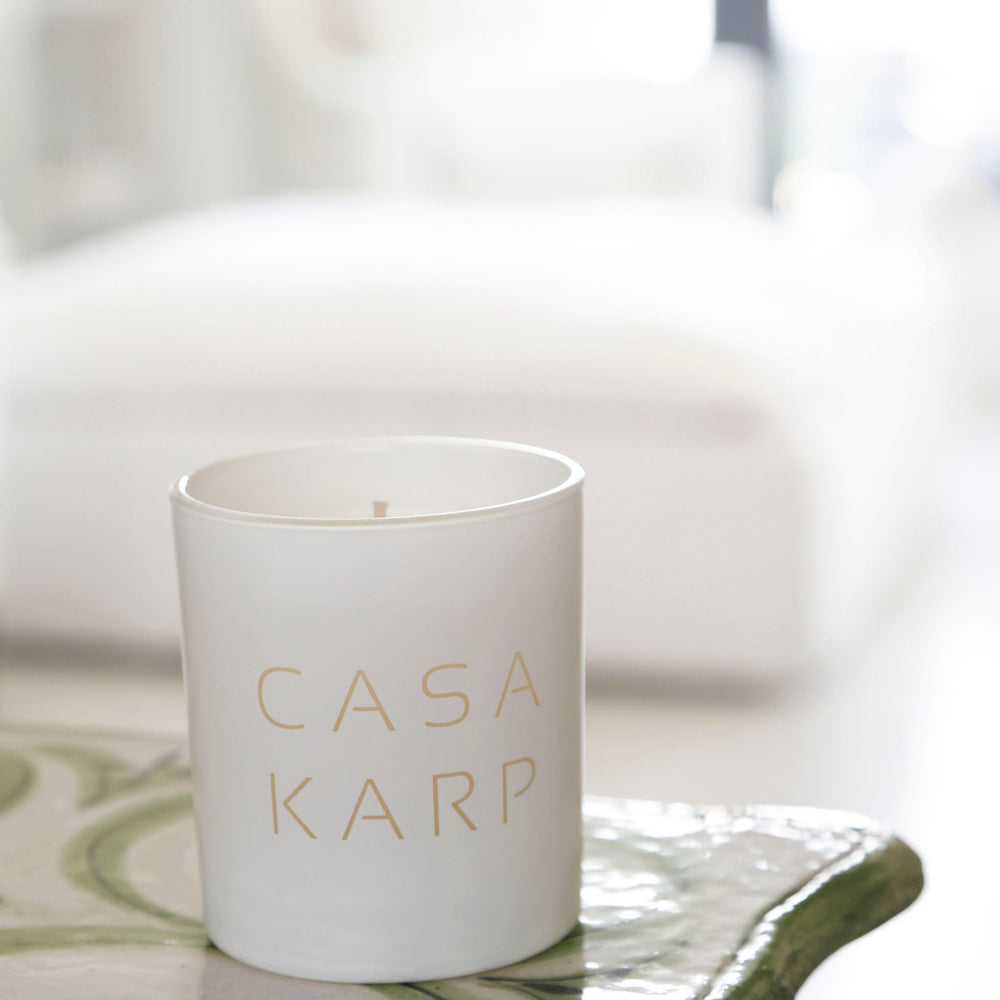 A black double-wick candle in an black glass jar with the brand name 'CASA FRANCO' engraved, set on a light beige surface with a blurred silver bowl in the background.