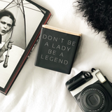 black dont be a lady be a legend candle next to a camera, book, and black pillow