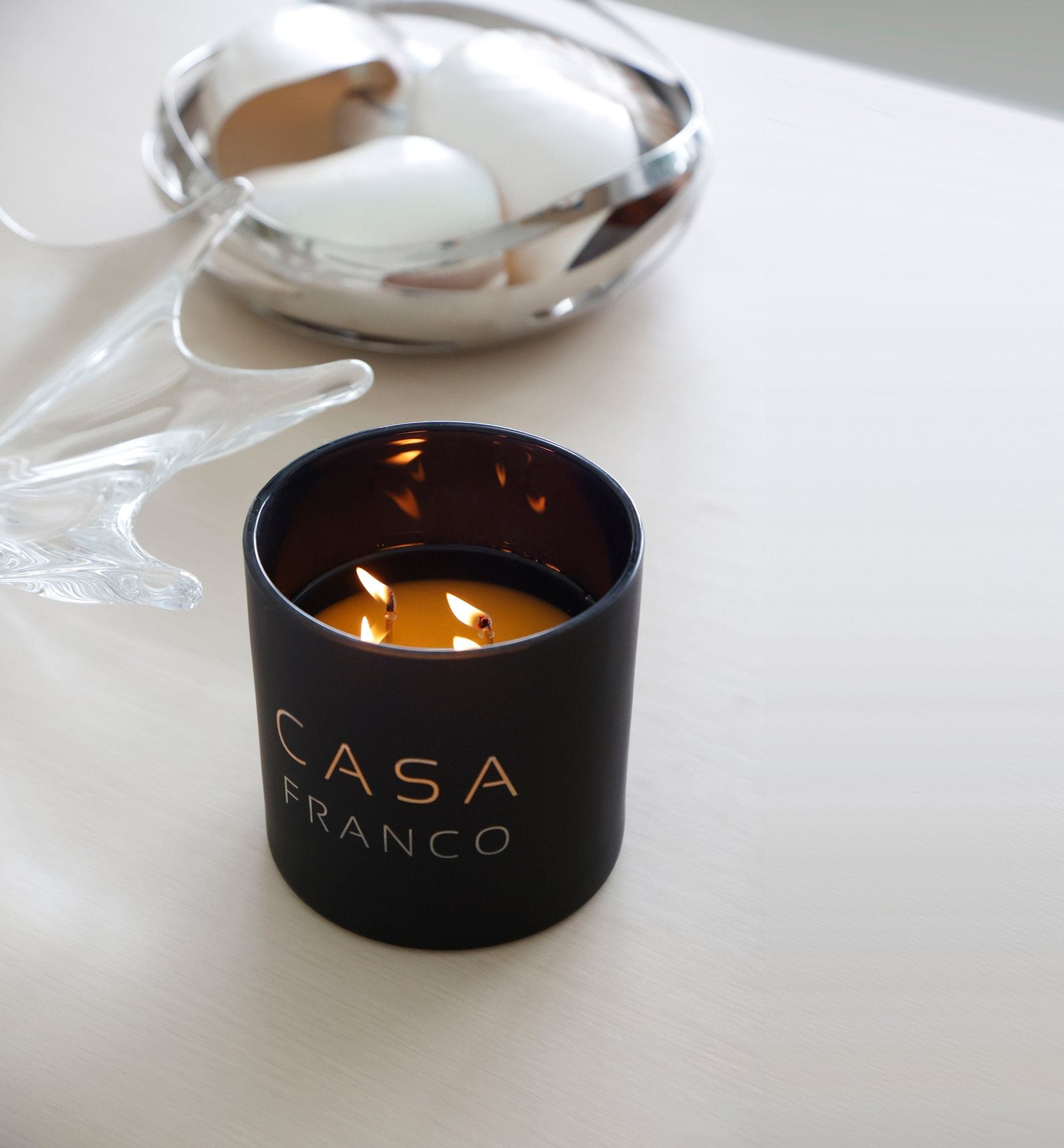 A black glass Casa cylinder candle, lit, on a white table beside a glass sculpture