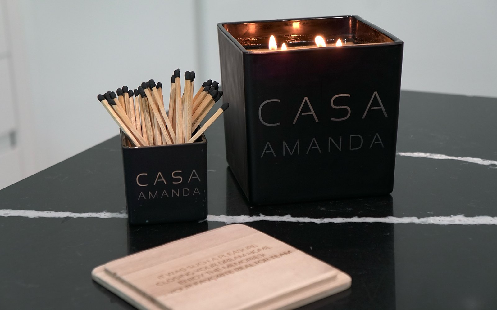 A lit black Casa candle engraved Casa Amanda, with its match holder and a wooden coaster