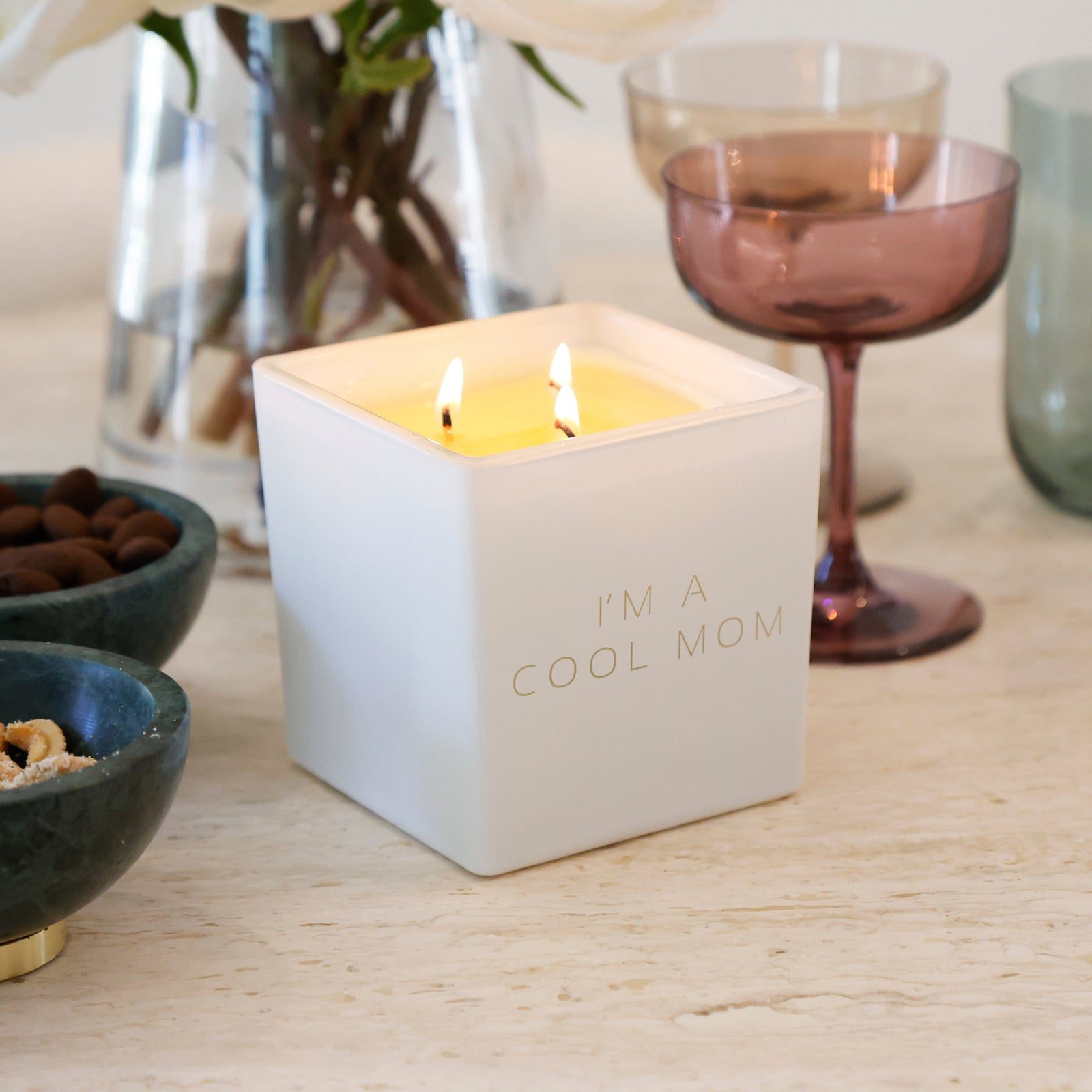 A lit white candle engraved I'm a cool mom on a travertine table beside blush coupe glasses